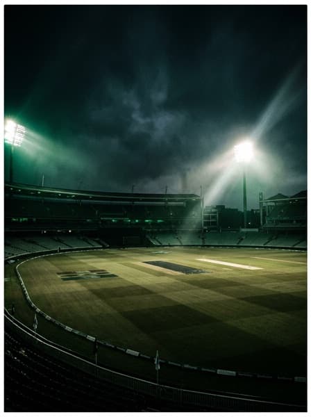 An empty floodlit cricket stadium at night, long shadows on the pitch